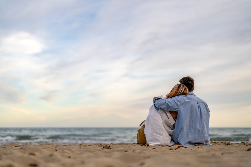 A couple sits closely together on a sandy beach, embracing as they look out at the ocean under a peaceful sky, symbolizing emotional connection and healing through couples therapy and marriage counseling. The calming setting represents support, communication, and strengthening relationships.