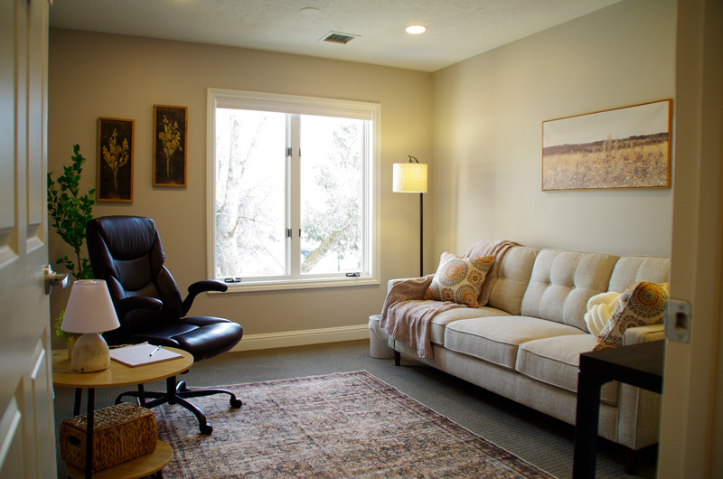A warm and inviting therapy room at Lighthouse Trauma Therapy, featuring a beige tufted sofa with decorative pillows and a soft throw blanket, a black leather recliner, a wooden side table with a lamp and notebook, and a large window allowing natural light to fill the space. The room is decorated with nature-inspired artwork, a floor lamp, and a patterned area rug for a calming atmosphere.