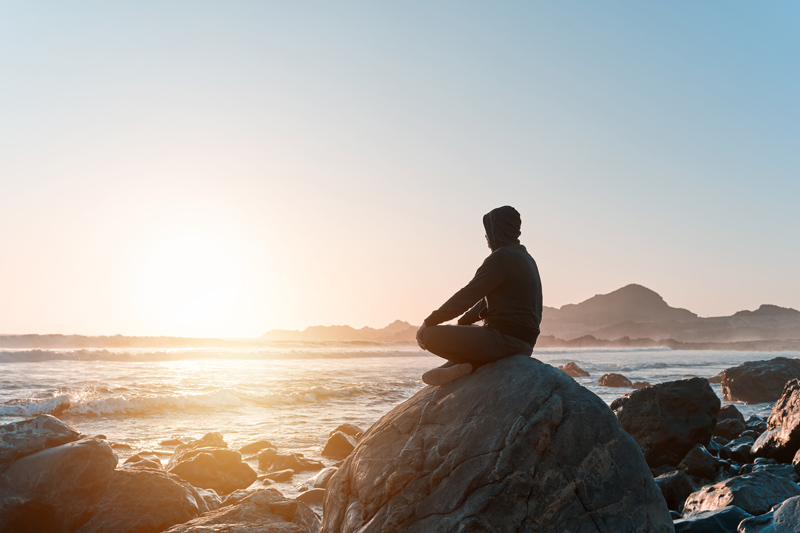 A person sits alone on a large rock by the ocean, gazing at the sunrise in a moment of quiet reflection, symbolizing healing and self-discovery through individual therapy. The peaceful coastal setting represents mindfulness, personal growth, and emotional well-being.