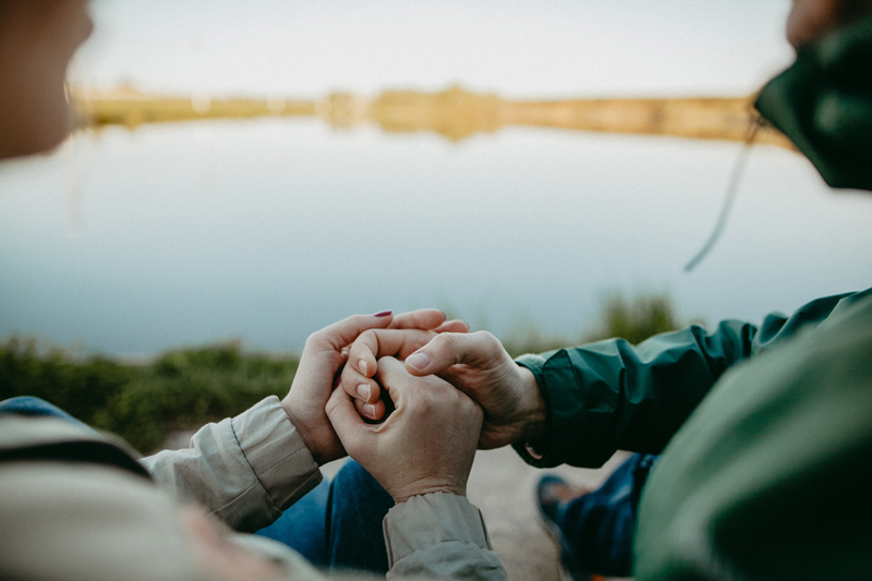 A couple sits near a calm lake, holding hands in a moment of emotional connection, symbolizing healing and reconciliation through marriage counseling. The peaceful natural setting represents trust, communication, and strengthening relationships.