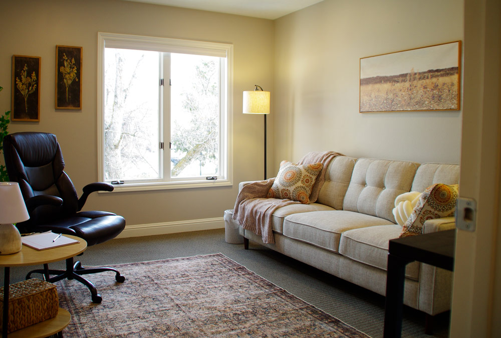 A warm and inviting therapist's office featuring a beige tufted sofa with decorative pillows and a soft throw blanket, a black leather swivel chair, and a wooden side table with a lamp and notepad. The room has a large window allowing natural light, a floor lamp, framed wall art, and a textured area rug on a neutral carpeted floor.