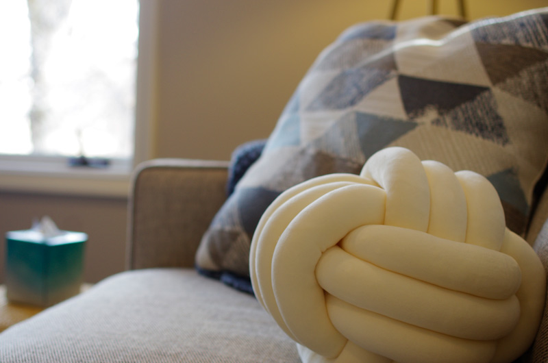 A-close-up-of-a-comfortable-couch-at-lighthouse-trauma-therapy,-featuring-a-unique-knot-shaped-cushion,-a-geometric-patterned-pillow,-and-a-window-in-the-background,-enhancing-the-room’s-cozy-atmosphere.