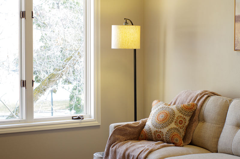 A-detailed-view-of-a-therapy-room-at-lighthouse-trauma-therapy,-featuring-a-soft-beige-couch-with-patterned-pillows,-a-cozy-blanket,-a-floor-lamp,-and-a-large-window-with-a-view-of-outside-trees,-enhancing-the-peaceful-atmosphere.