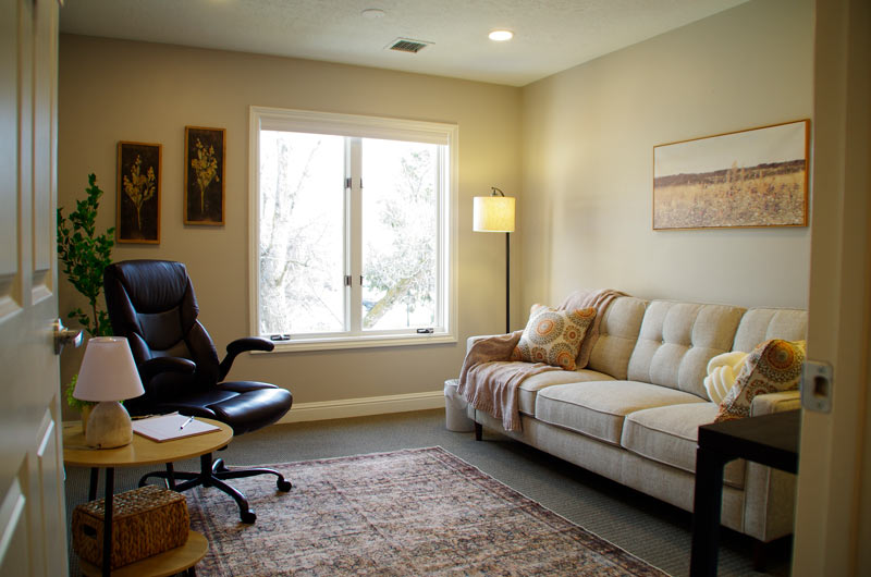 A-comfortable-therapy-room-at-lighthouse-trauma-therapy-with-a-large-window,-natural-light,-a-beige-couch-with-decorative-pillows,-a-black-leather-chair,-and-a-floor-lamp,-creating-a-warm-and-soothing-environment.