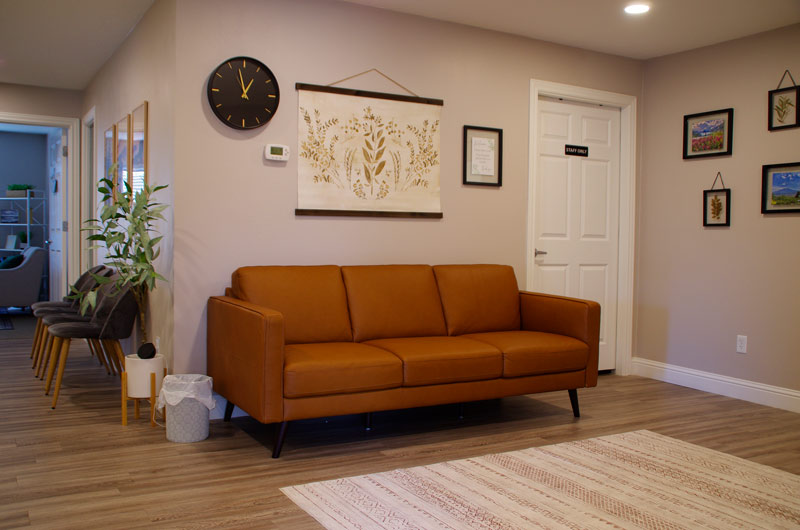 A-modern-waiting-room-at-lighthouse-trauma-therapy-featuring-a-brown-leather-couch,-wall-art,-and-framed-pictures-on-a-neutral-colored-wall.-The-room-has-wooden-flooring,-a-round-wall-clock,-and-a-warm,-inviting-ambiance.