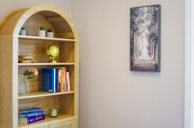 A-therapy-room-at-lighthouse-trauma-therapy-featuring-a-light-wood-bookshelf-with-books,-small-plants,-a-globe,-and-a-candle,-alongside-a-framed-painting-of-a-tree-lined-path,-creating-a-serene-and-thoughtful-environment.